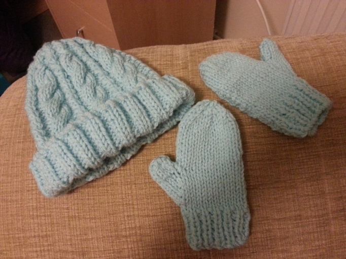 Pale blue cable hat and plain mittens laid side by side